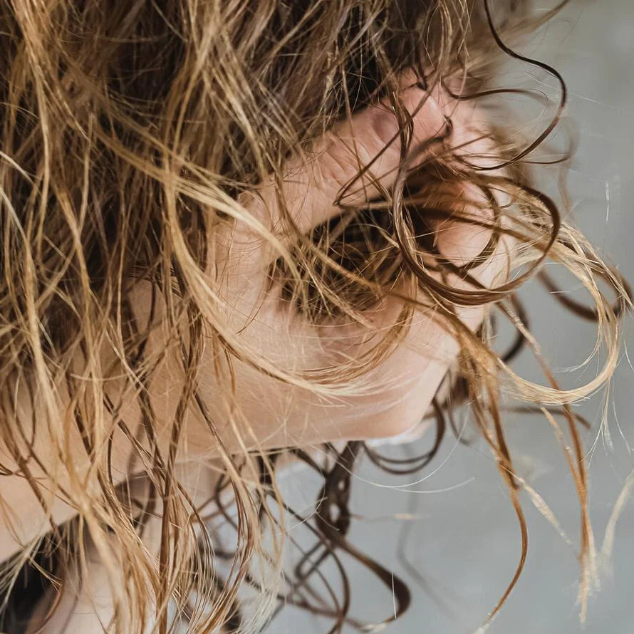 A square, close-up photo of a young Irish woman with a frustrated and unhappy expression. Her gaze is directed at the camera. She has wavy, brown hair with visibly greasy roots and dry, frizzy ends. The background is a plain, neutral-colored studio wall.