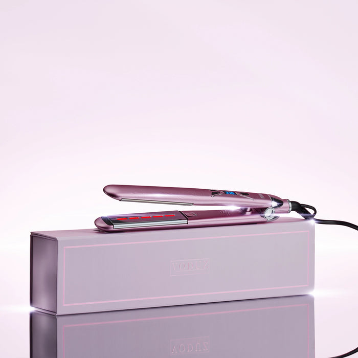 A pink Voduz Infrared Straightener hair straightener resting on top of its matching pink product box, set against a clean light pink studio background with a reflective surface below.