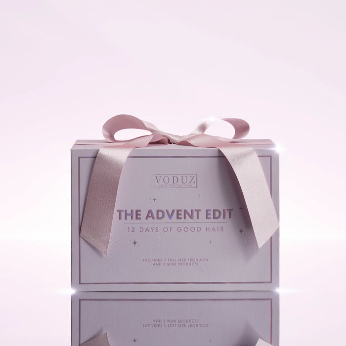 A square gift box for VODUZ The Advent Edit hair care set, wrapped with a pale pink satin ribbon bow, set against a soft light pink background with a reflection below.