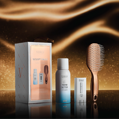 Voduz Sculpt Vault hair product set with box, spray bottle, and brush on a reflective surface with a warm glow.